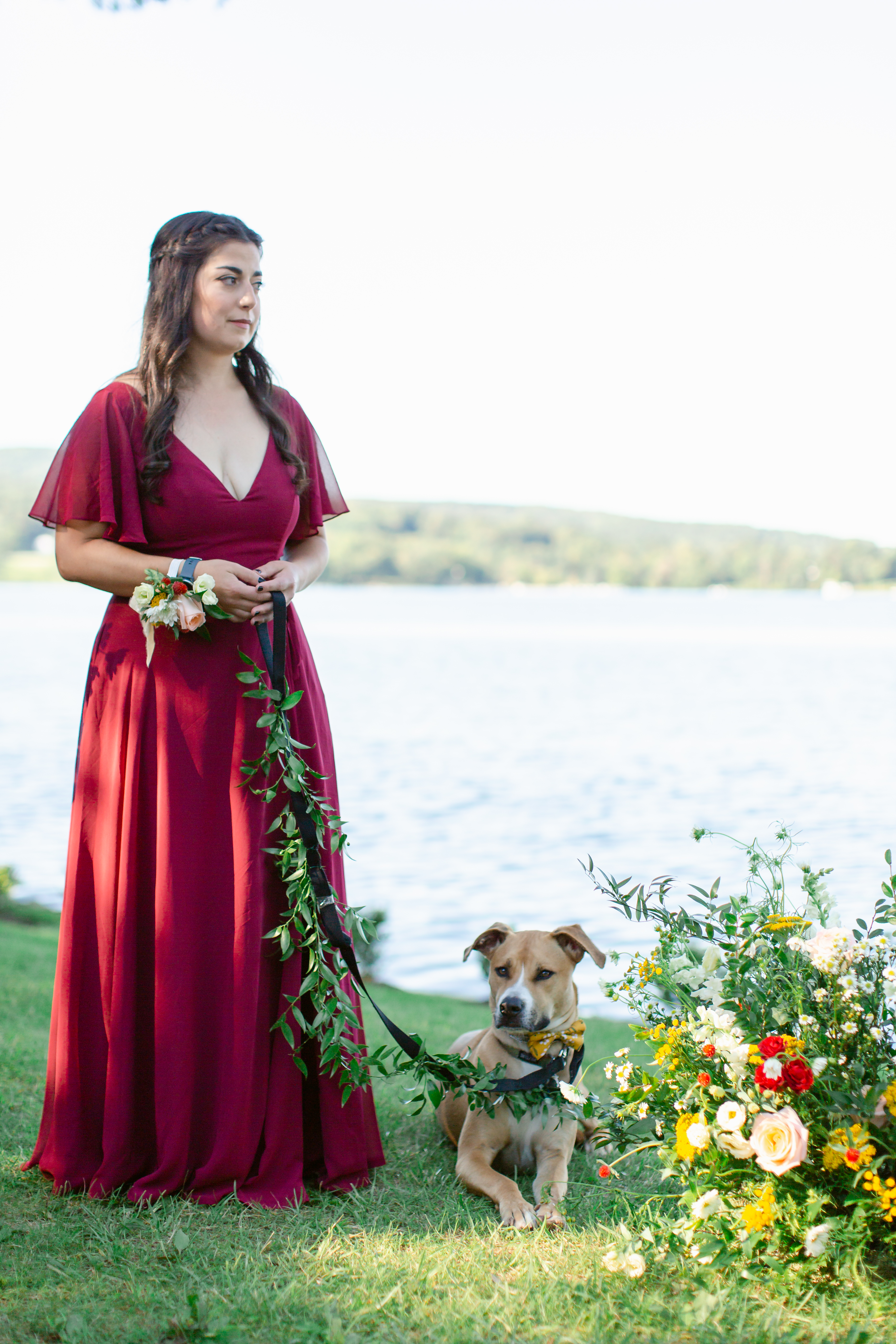woman in a red dress holding a leash with a dog by the side of a lake with a bouquet of flowers next tot he dog